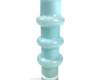May include: A tall, cylindrical glass vase in a pale, translucent light blue color. The vase features three distinct, rounded horizontal ridges or rings stacked vertically along its body, with a smooth, clear glass base at the bottom.