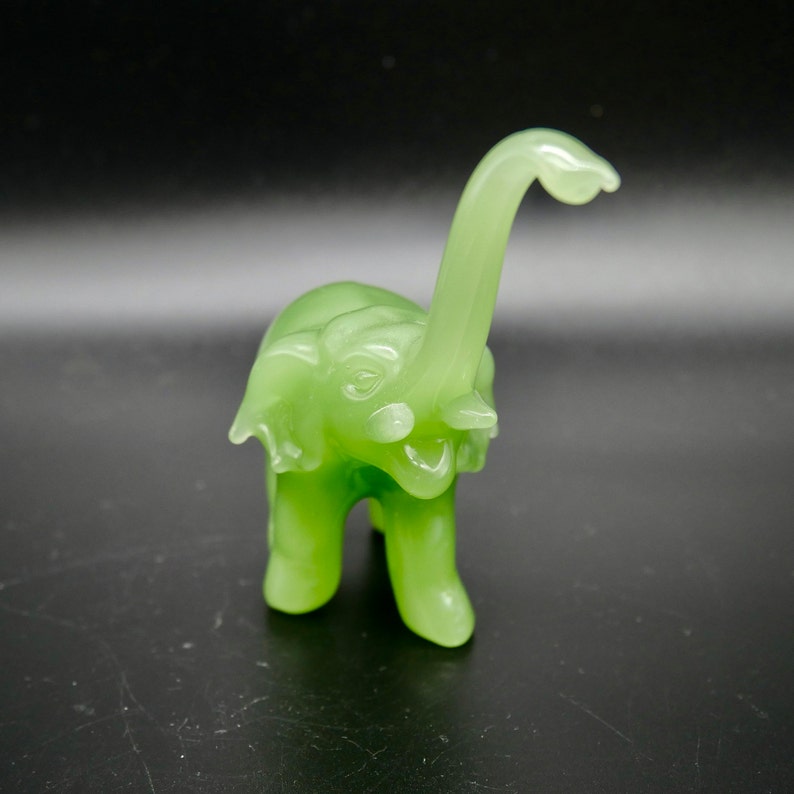 K&ouml;nnte beinhalten: Eine kleine, hellgr&uuml;ne Elefantenfigur mit erhobenem R&uuml;ssel. Der Elefant hat einen l&auml;chelnden Ausdruck und besteht aus einem durchscheinenden Material. Die Figur ist ein Dekorationsartikel, wahrscheinlich f&uuml;r ein Regal oder einen Schreibtisch.