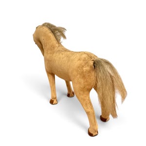 May include: A tan plush horse toy with a textured fabric body, a light-colored synthetic mane, and a matching long tail. The figure features visible stitched seams along the back and neck, with dark brown accents on the lower legs and hooves.