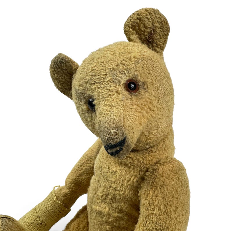 Pode incluir: Um urso de peluche de estilo vintage feito de tecido mohair texturizado castanho claro. O urso apresenta olhos de vidro castanho escuro, um nariz e boca costurados a preto e orelhas arredondadas. O pelo mostra sinais de desgaste.