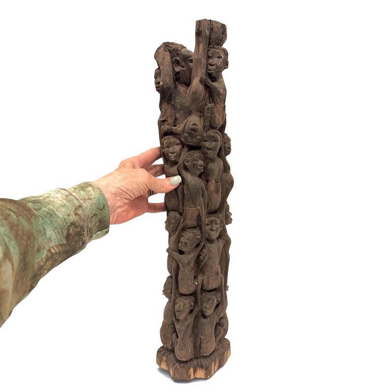 May include: A tall, dark brown wooden sculpture carved from a single piece of wood, featuring a vertical stack of numerous human figures intertwined and climbing upwards, with detailed facial expressions and limbs visible throughout the entire structure.