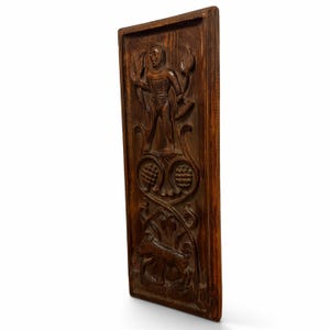 May include: A rectangular dark brown wooden wall plaque featuring a hand-carved relief. The top section depicts a standing figure holding a tool, while the lower section shows a small animal amidst scrolling floral patterns and circular motifs.