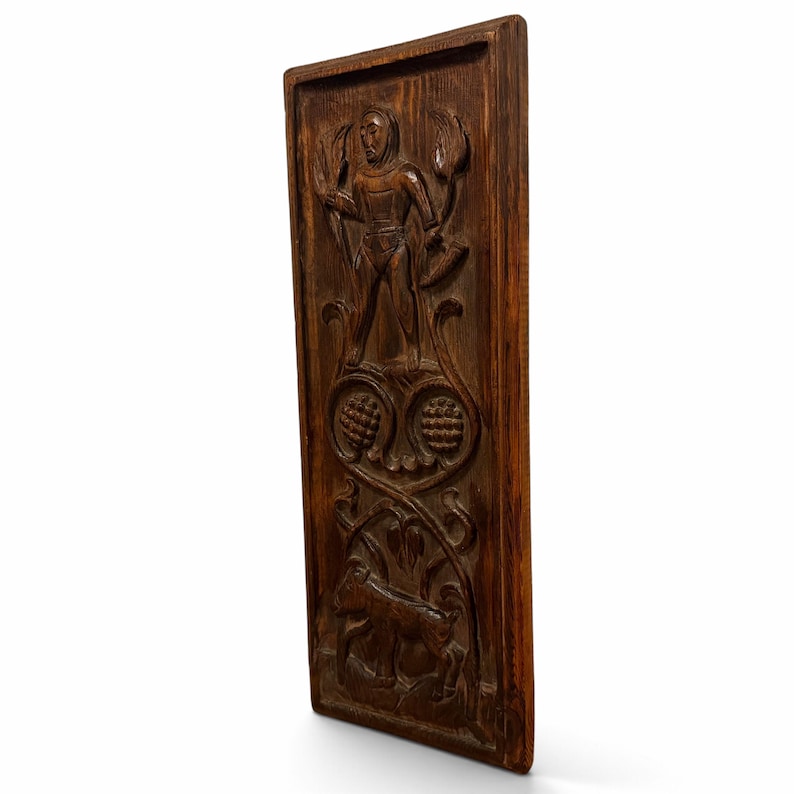 May include: A rectangular dark brown wooden wall plaque featuring a hand-carved relief. The top section depicts a standing figure holding a tool, while the lower section shows a small animal amidst scrolling floral patterns and circular motifs.