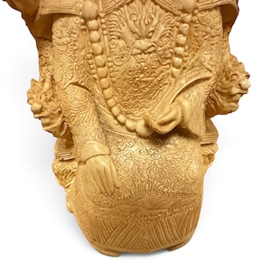 Op de afbeelding: Een cr&egrave;mekleurig uitgesneden beeldje met ingewikkelde reli&euml;fpatronen van draken en wolken op de kleding. Het figuur draagt een lange kralenketting en houdt een waaier in de hand, met twee gebeeldhouwde koppen van mythische wezens aan de zijkanten.