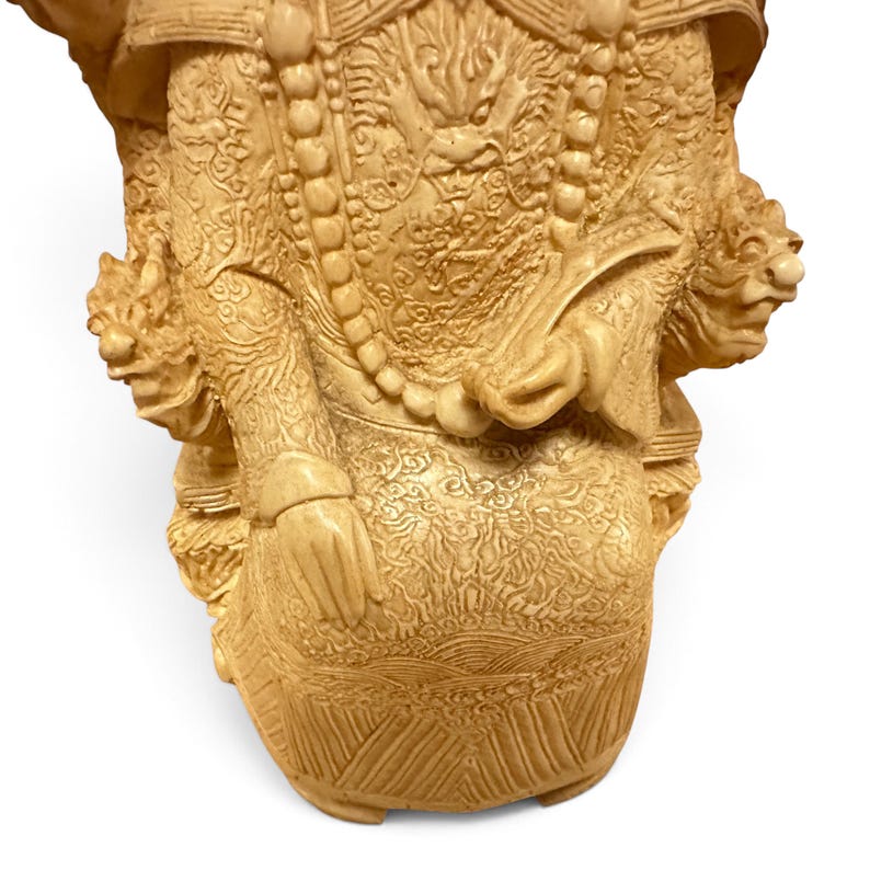 Op de afbeelding: Een cr&egrave;mekleurig uitgesneden beeldje met ingewikkelde reli&euml;fpatronen van draken en wolken op de kleding. Het figuur draagt een lange kralenketting en houdt een waaier in de hand, met twee gebeeldhouwde koppen van mythische wezens aan de zijkanten.