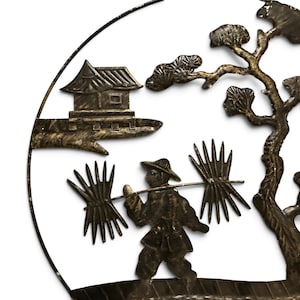 May include: A circular metal wall art piece featuring a dark bronze finish. The design depicts a figure wearing a hat, carrying two bundles of sticks on a pole across their shoulders, a small house on a ledge, and a tree within a circular frame.