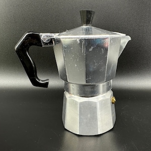 Puede incluir: Una cafetera de aluminio plateado con un mango de pl&aacute;stico negro. La cafetera tiene un cuerpo redondeado y un pico en el lateral.