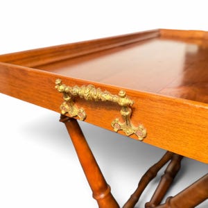 Puede incluir: Una bandeja de servicio rectangular de madera con un acabado liso y pulido y un borde elevado. La bandeja cuenta con asas met&aacute;licas de color dorado ornamentadas con detalles intrincados en ambos lados y descansa sobre una base de madera torneada.