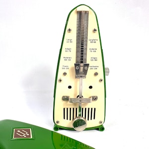 May include: A green and cream mechanical metronome featuring a central metal pendulum and a vertical scale with tempo markings including Largo, Adagio, Moderato, Presto, Larghetto, Andante, Allegro, and Prestissimo. A matching green cover is in the foreground.