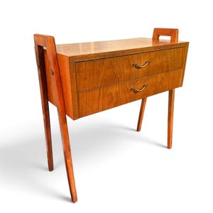 Op de afbeelding: Een houten bijzettafel met een rechthoekig blad en twee lades. Het meubel heeft een lichtbruine houtnerfafwerking, gebogen poten en twee kleine messingkleurige metalen handgrepen op elke lade. Het frame bevat verticale steunen.