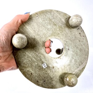 May include: A round, light green stone object with three spherical feet and a central hole. The object has a textured surface and a small sticker that reads "MADE IN INDIA".