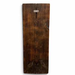 May include: A rectangular dark brown wooden board with a visible wood grain pattern and natural knots. A small, rectangular silver-colored metal mounting bracket is attached to the upper center of the board for hanging purposes.