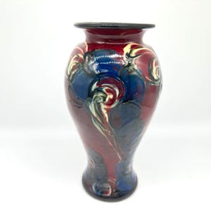 May include: A decorative ceramic vase with a dark red base and a black rim. The vase features swirling patterns in blue, cream, and red. The vase has a bulbous shape and a narrow neck, suitable for floral arrangements.