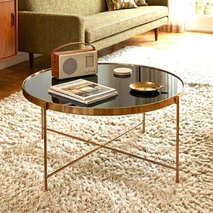 Puede incluir: Una mesa de centro redonda con tapa de cristal oscuro y estructura de metal dorado con patas cruzadas. Sobre la mesa hay una radio rectangular de color beige con asa, una pila de libros, un posavasos circular pequeño y un plato metálico dorado.