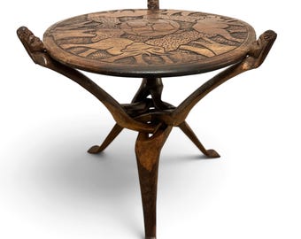 May include: A round wooden side table featuring a dark brown finish. The tabletop is intricately carved with animal figures. The base consists of three legs shaped like human figures, with their heads supporting the table edge.