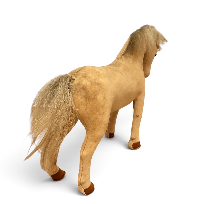 May include: A tan plush horse toy with a textured fabric body, a long light-colored mane, and a matching tail. The horse features dark brown accents on the lower legs and hooves, with visible stitched seams running along the back and legs.