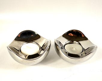 Puede incluir: Dos portavelas de té de metal color plata con una forma curva, lisa y reflectante similar a un cuenco. Cada portavelas presenta una hendidura central circular diseñada para sostener una pequeña vela blanca redonda.
