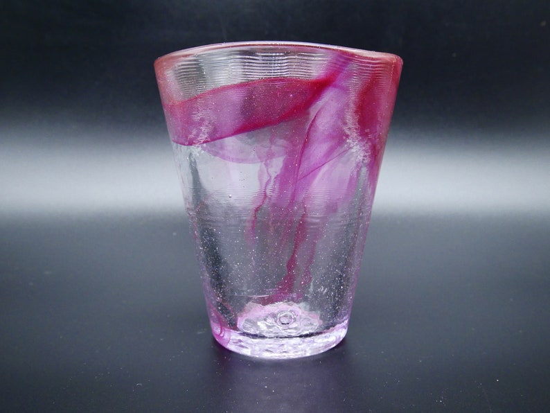 Puede incluir: Un vaso de vidrio transparente con forma acampanada y superficie texturizada. El vidrio presenta un dise&ntilde;o vibrante y arremolinado de tonos magenta y rosa en el interior, creando un efecto visual &uacute;nico. La base es de un tono rosa m&aacute;s claro.