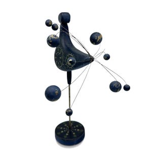 May include: A dark blue wooden bird sculpture mounted on a vertical metal rod and a circular base. The bird features a painted pattern of light green dots and thin wire extensions radiating outward, each tipped with a small dark blue sphere.