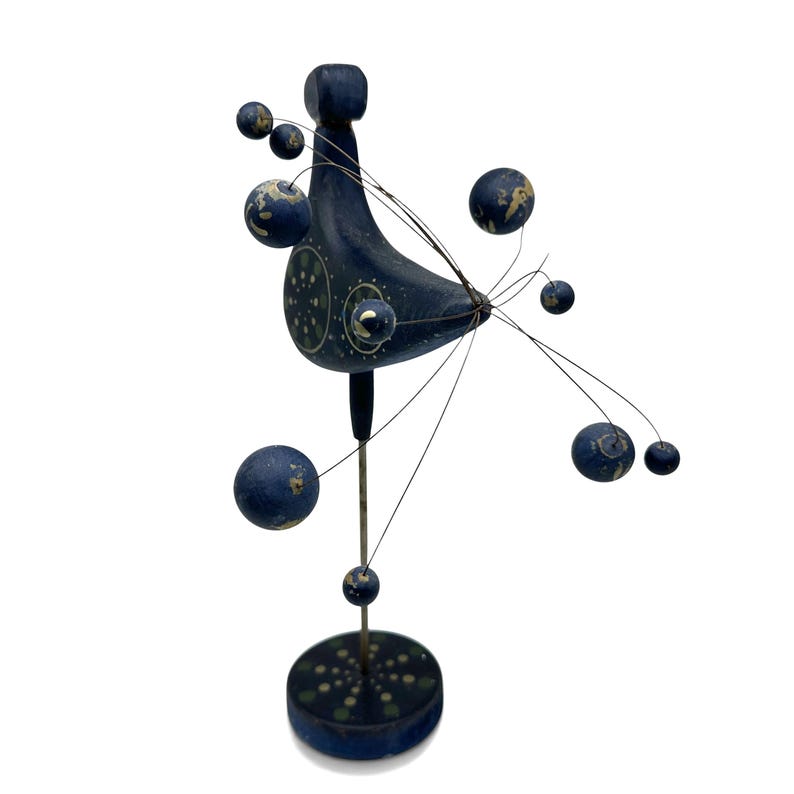 May include: A dark blue wooden bird sculpture mounted on a vertical metal rod and a circular base. The bird features a painted pattern of light green dots and thin wire extensions radiating outward, each tipped with a small dark blue sphere.