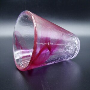 Puede incluir: Un vaso de vidrio transparente con un interior y borde magenta vibrante. El vidrio tiene una apariencia texturizada, ligeramente esmerilada, con estr&iacute;as verticales en la parte inferior. El color magenta se arremolina y se mezcla dentro del vidrio.