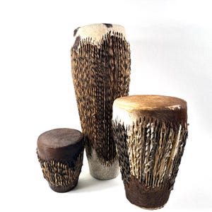 Può includere: Un set di tre tamburi con pelli animali e corpi avvolti in materiale fibroso testurizzato marrone scuro e marrone chiaro. I tamburi variano in altezza e larghezza, presentando motivi di pelliccia naturale sulle superfici superiori.