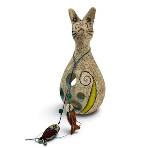 May include: A speckled beige ceramic cat figurine with a teardrop-shaped body, featuring cut-out circular holes, a yellow crescent accent, and a dark string hanging from its neck with two small, colorful ceramic fish charms attached.