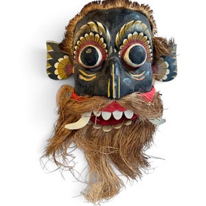 Könnte beinhalten: Eine handgeschnitzte Holzmaske mit schwarzem Gesicht, großen runden Augen, geschwungenen Augenbrauen und spitzen Ohren. Die Maske hat einen roten Mund mit weißen Zähnen, markanten gebogenen Fangzähnen und einen dichten, faserigen braunen Bart.