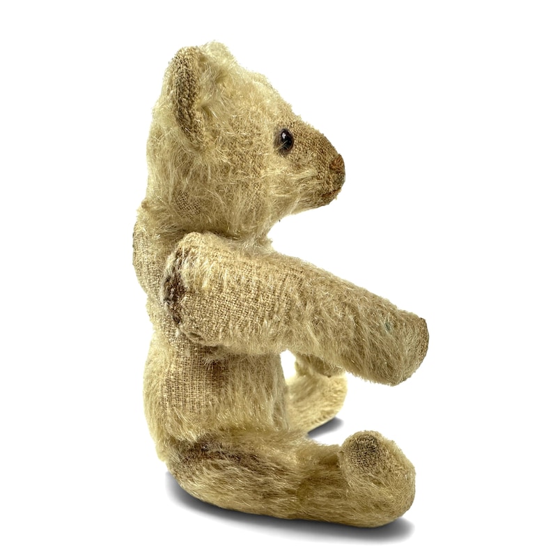 May include: A side profile of a small, vintage-style teddy bear crafted from textured, light beige mohair fabric. The bear features a dark brown stitched nose, small dark eyes, and visible seams along the limbs and torso, showing a worn appearance.