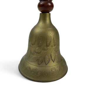 May include: A brass hand bell featuring an etched leaf pattern on the exterior surface. The bell has a rounded, dark brown wooden handle attached to the top. The metal body displays a muted gold tone with visible engraved details throughout.