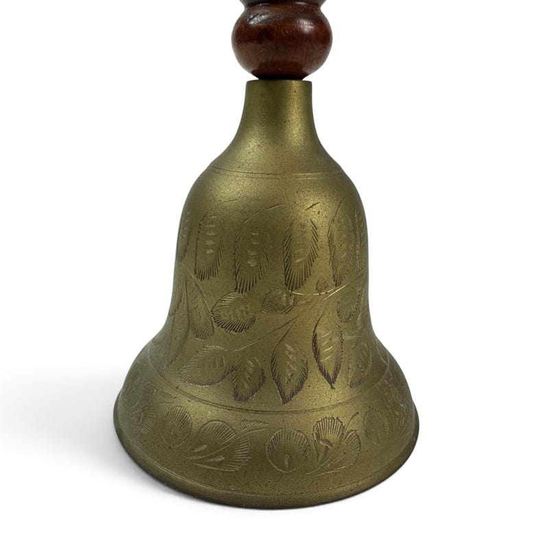 May include: A brass hand bell featuring an etched leaf pattern on the exterior surface. The bell has a rounded, dark brown wooden handle attached to the top. The metal body displays a muted gold tone with visible engraved details throughout.