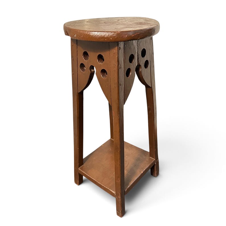 May include: A brown wooden plant stand featuring a round top, four straight legs, and a square lower shelf. The upper section of the stand displays decorative cutouts with circular holes, creating a distinct architectural pattern.