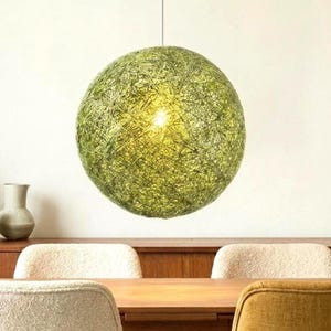 May include: A spherical pendant light with a woven, green exterior. The light hangs from a single cord and emits a warm glow. The fixture is suspended above a wooden table and chairs, creating a cozy atmosphere.
