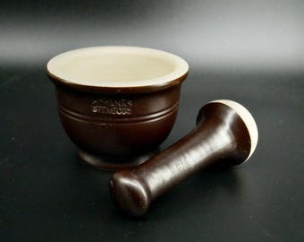 May include: A dark brown ceramic mortar and pestle set with a cream-colored interior. The mortar features the embossed text HÖGANÄS STENGODS on the side. The pestle has a matching dark brown handle and a cream-colored grinding end.