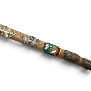Puede incluir: Un bast&oacute;n de senderismo de madera decorado con varias insignias met&aacute;licas de recuerdo. Las insignias presentan diversos dise&ntilde;os, incluyendo una cabra mont&eacute;s, un escudo con el texto INNSBRUCK, un paisaje de monta&ntilde;a verde y otros emblemas.