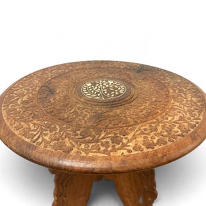 May include: A round wooden stool featuring intricate floral carvings across the surface and a central circular inlay depicting a stylized tree with white leaves. The stool has a natural brown finish and carved legs visible at the base.