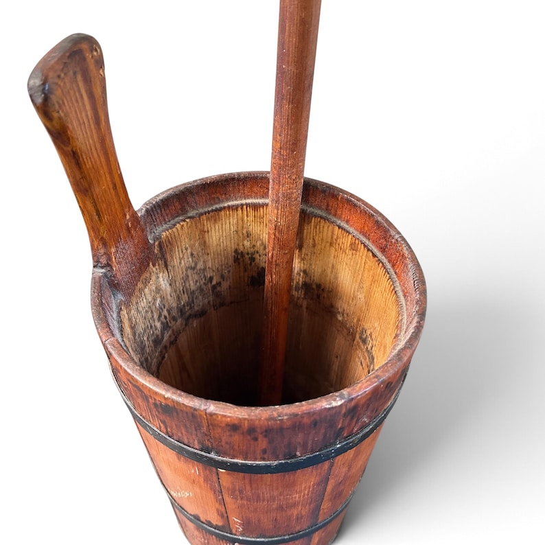 May include: A tall, cylindrical wooden butter churn featuring a weathered brown finish, dark metal bands encircling the exterior, a vertical wooden plunger handle extending from the center, and a curved wooden side handle attached to the rim.