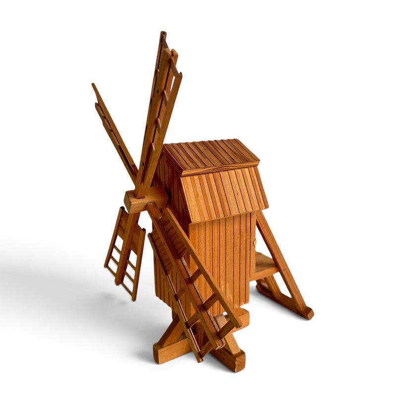 May include: A wooden windmill model featuring a vertical rectangular body with a sloped roof, four lattice-style blades attached to a central hub, and a sturdy base structure with angled support beams, all constructed from light brown natural wood.