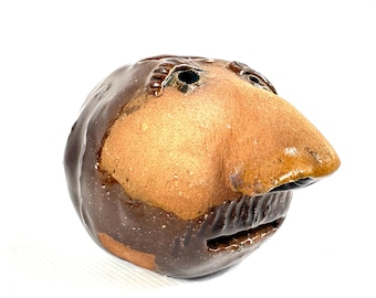 May include: A round, brown ceramic sculpture shaped like a human head with a large, prominent nose and a wide, open mouth. The surface features a glossy dark brown glaze on the sides and back, with a matte, textured tan finish on the face.