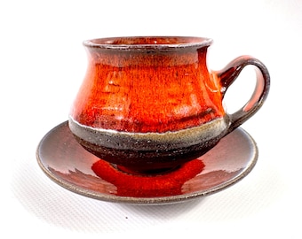 Puede incluir: Un juego de taza y platillo de café de cerámica hecho a mano. La taza y el platillo están esmaltados en tonos de rojo, marrón y negro. La taza tiene una forma redondeada con un asa. El platillo es redondo y complementa el diseño de la taza.