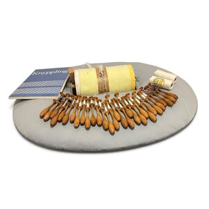 May include: A lace making pillow with a yellow cylindrical drum, numerous wooden bobbins with white thread, and a blue instructional book titled Knyppling. Two spools of white thread are positioned on the right side of the gray oval pillow.