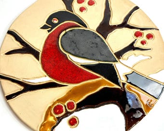 May include: A circular wall plaque featuring a glossy, raised-relief design of a bullfinch perched on a dark brown branch. The bird has a vibrant red chest, black head, and grey wings. Small red berries are scattered on the branches against a cream background.