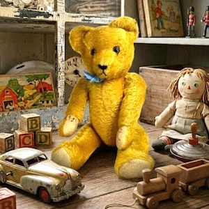May include: A vintage yellow teddy bear with a blue bow tie sits among other antique toys. A toy car, wooden train, and a rag doll are also visible. Wooden alphabet blocks and a vintage clock are in the background. The toys are arranged on a wooden surface.