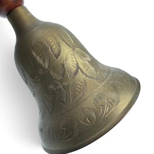 May include: A brass hand bell featuring an etched leaf pattern across its surface. The bell has a flared opening and is attached to a smooth, rounded wooden handle at the top. The metal has a muted, aged finish with visible engraved details.