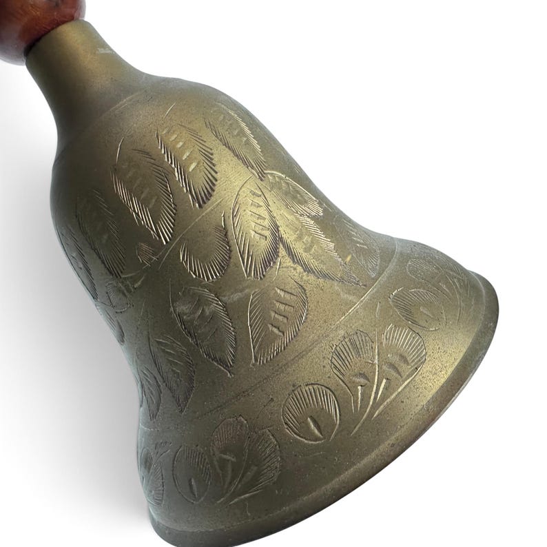May include: A brass hand bell featuring an etched leaf pattern across its surface. The bell has a flared opening and is attached to a smooth, rounded wooden handle at the top. The metal has a muted, aged finish with visible engraved details.