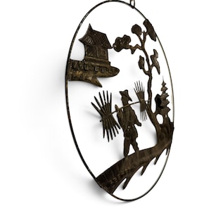 May include: A circular dark metal wall art piece featuring a silhouette scene of a person carrying bundles on a pole across their shoulders. The design includes a small building, a tree, and a pagoda, all enclosed within a thin metal ring frame.