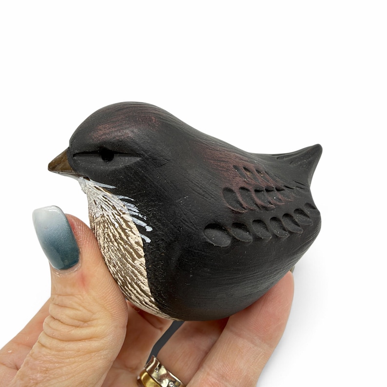 May include: A small, hand-carved wooden bird figurine featuring a dark brown and black finish. The bird has a pointed beak, carved wing details, and a textured, light-colored chest area with white painted accents on the front.