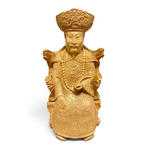 Op de afbeelding: Een lichtbruin uitgesneden beeldje van een zittende figuur met een versierde hoed en traditionele gewaden met complexe drakenmotieven. De figuur heeft een lange baard, houdt een gebogen voorwerp vast en zit op een stoel met drakenkoppen.