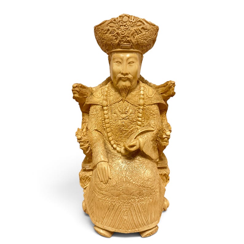 Op de afbeelding: Een lichtbruin uitgesneden beeldje van een zittende figuur met een versierde hoed en traditionele gewaden met complexe drakenmotieven. De figuur heeft een lange baard, houdt een gebogen voorwerp vast en zit op een stoel met drakenkoppen.