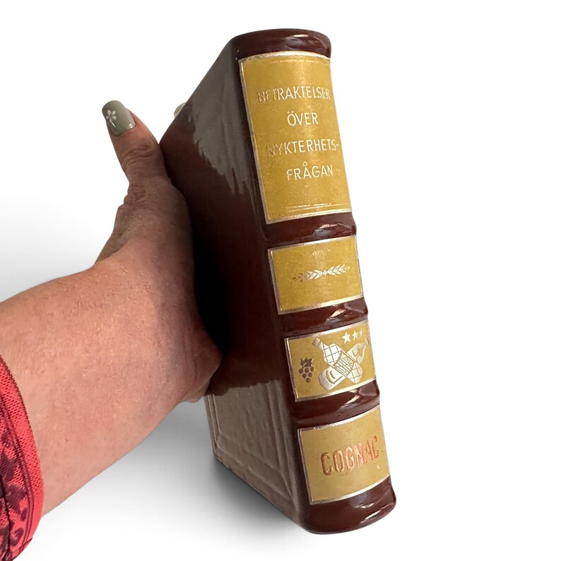 Puede incluir: Un decantador de cer&aacute;mica marr&oacute;n con forma de libro antiguo y acabado brillante. El lomo presenta etiquetas doradas con el texto "BETRAKTELSER &Ouml;VER NYKTERHETSFR&Aring;GAN" y "COGNAC", junto con ilustraciones de un racimo de uvas y botellas.